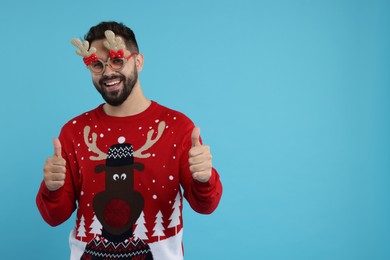 Photo of Happy young man in Christmas sweater and funny glasses showing thumbs up on light blue background. Space for text