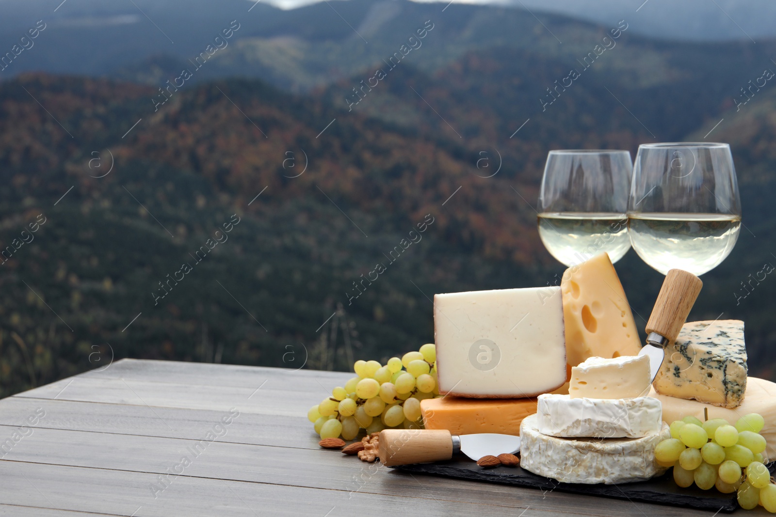 Photo of Different types of delicious cheeses, snacks and wine on wooden table against mountain landscape. Space for text