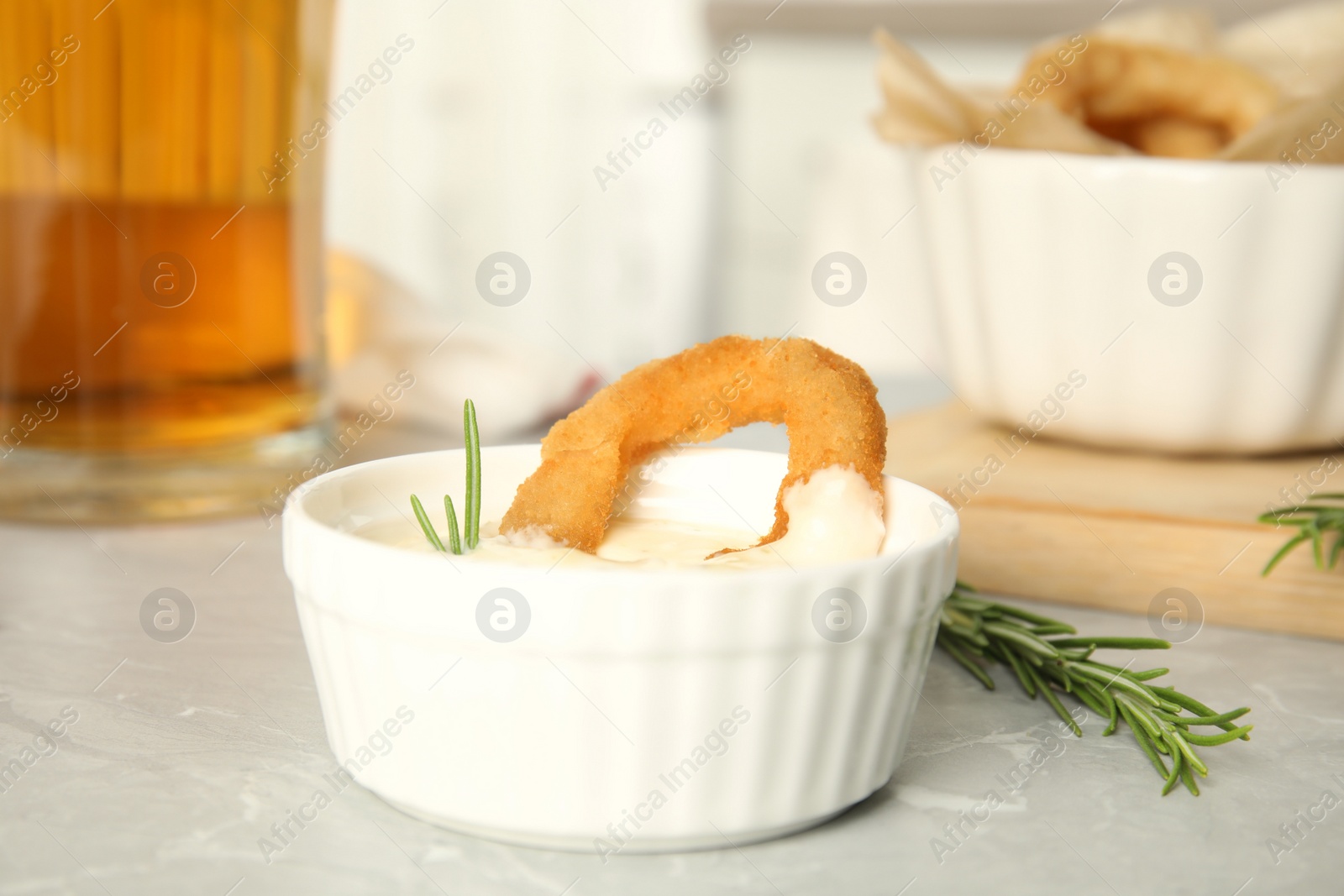 Photo of Delicious crunchy fried onion ring in bowl with sauce on grey marble table