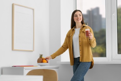Beautiful young woman with delicious smoothie taking fruit at home. Space for text