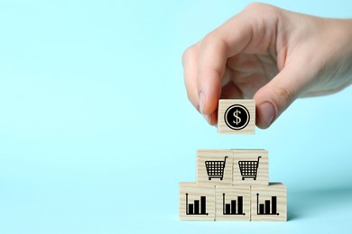 Image of Woman putting cube with dollar sign on other ones with charts and shopping carts against light blue background, closeup. Space for text