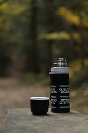 Black thermos and cup lid on wooden bench outdoors, space for text