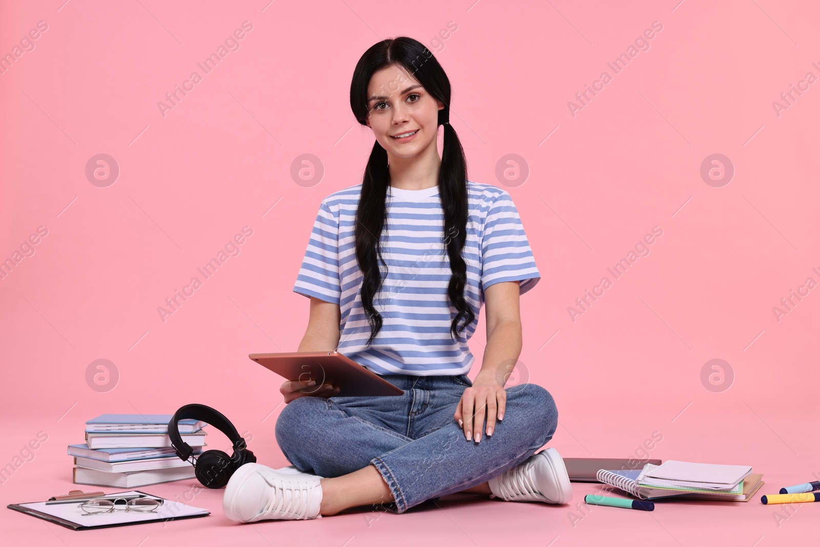 Photo of Smiling student with tablet sitting among books and stationery on pink background