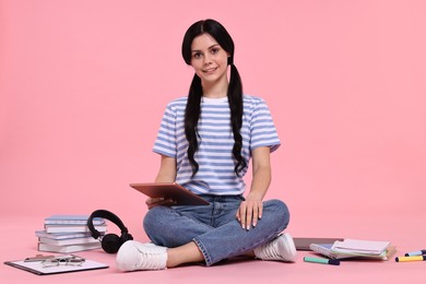 Smiling student with tablet sitting among books and stationery on pink background