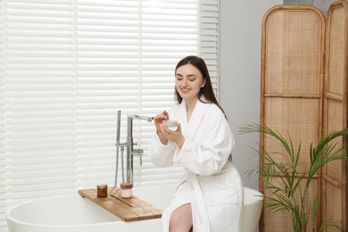 Photo of Beautiful woman in white robe applying cream in bathroom