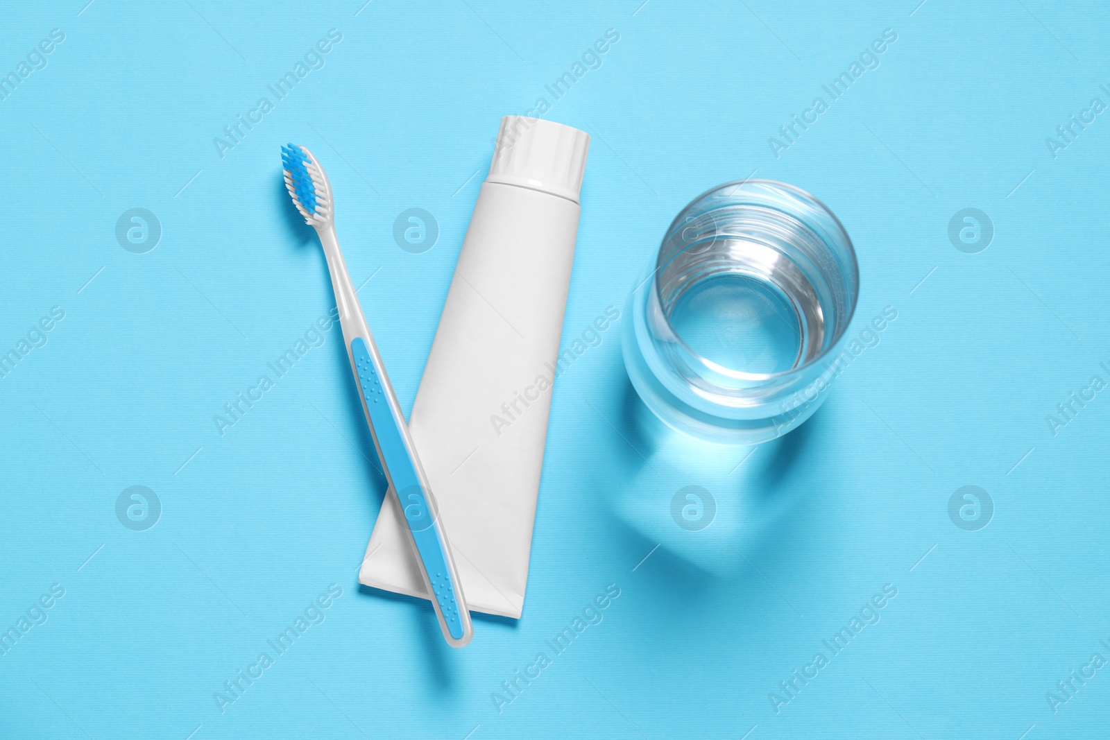 Photo of Plastic toothbrush with paste and glass of water on light blue background, flat lay