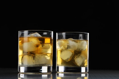 Tasty whiskey with ice in glasses on mirror table against black background, closeup