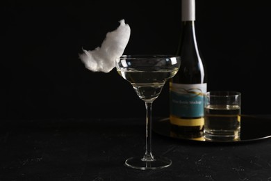 Photo of Cocktail with tasty cotton candy and bottle of alcohol drink on dark textured table against black background, space for text