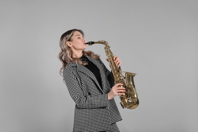 Beautiful young woman in elegant suit playing saxophone on grey background