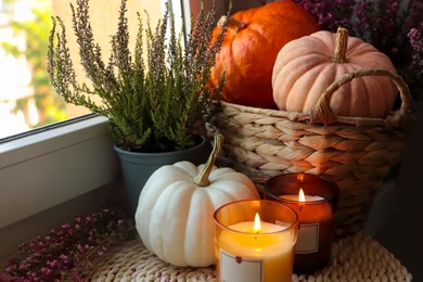 Photo of Beautiful heather flowers, burning candles and wicker basket with pumpkins near window indoors