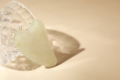 Photo of Jade gua sha tool near glass on beige background, space for text