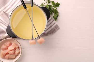 Fondue pot with oil, forks, raw meat pieces and parsley on white wooden table, flat lay. Space for text