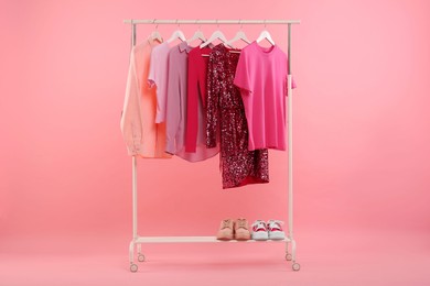 Rack with different stylish women`s clothes and sneakers on pink background