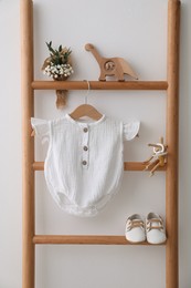 Photo of Baby bodysuit, shoes, toys and small bouquet on ladder near white wall