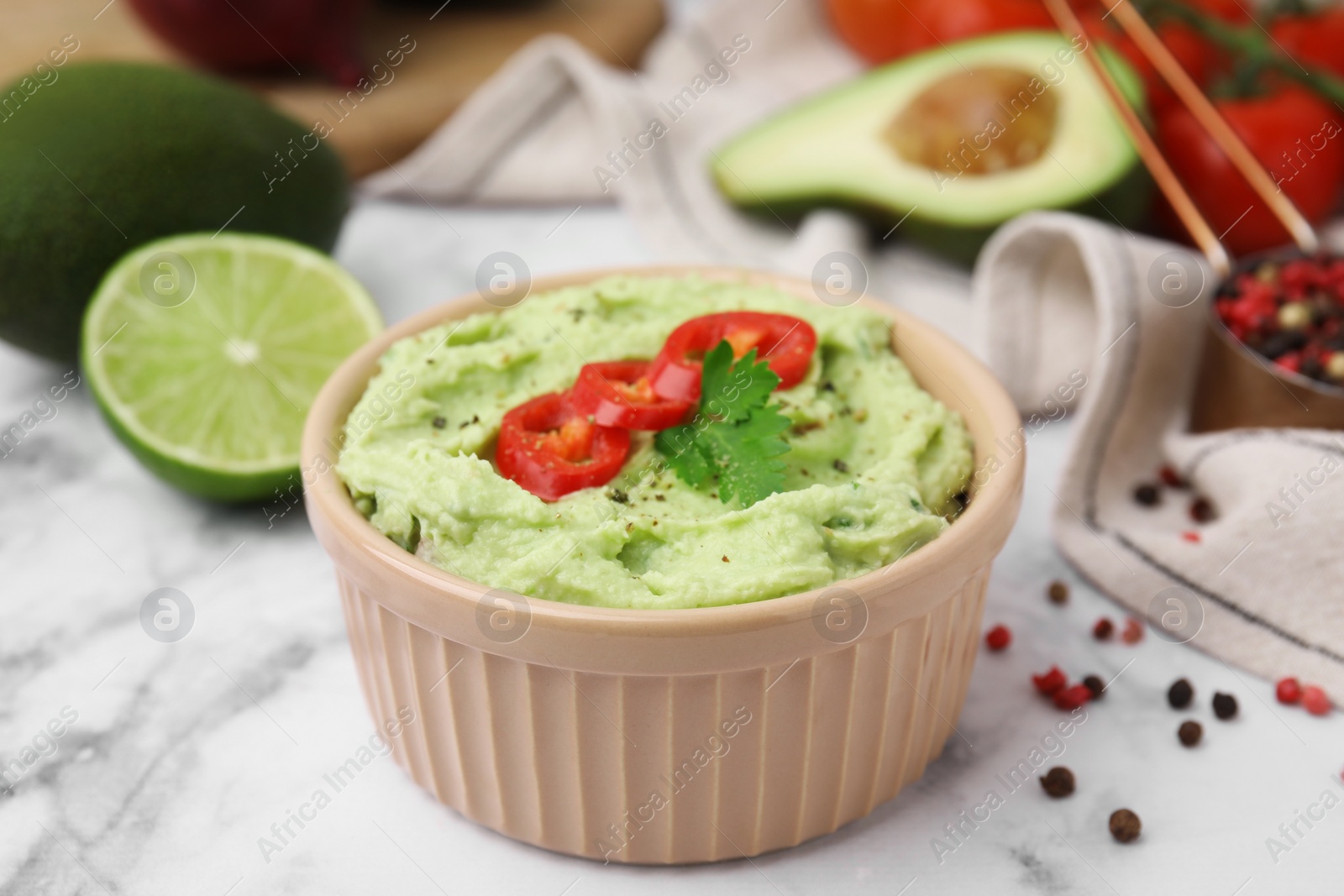 Photo of Bowl of delicious guacamole and ingredients on white marble table, closeup