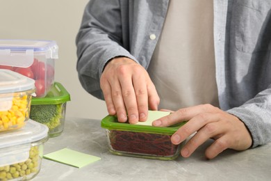 Photo of Man sticking paper note onto container with cut beets at light grey table, closeup. Food storage
