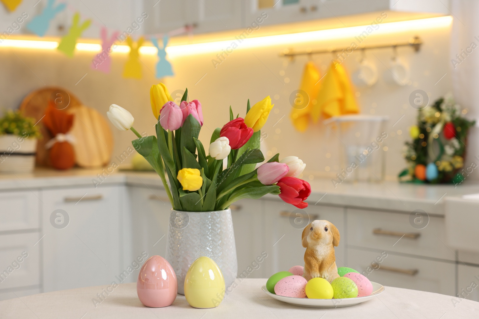 Photo of Bouquet of tulips, painted eggs and Easter decorations on white table in kitchen