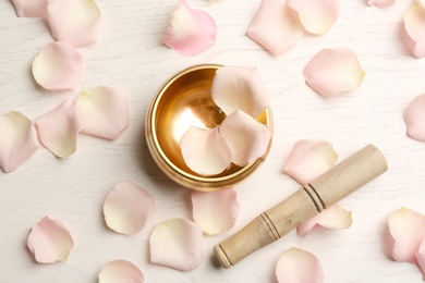 Photo of Golden singing bowl with petals and mallet on white wooden table, flat lay. Sound healing