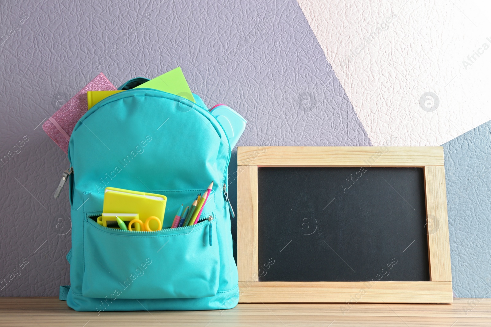 Photo of Backpack full of different school stationery and small blank chalkboard on table near color wall. Space for text