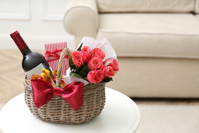 Photo of Wicker basket with gift, bouquet and wine on white table indoors. Space for text