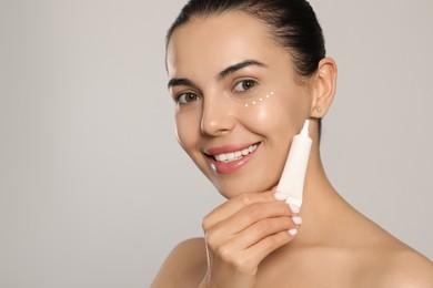 Beautiful young woman with cream on skin under eye against light grey background