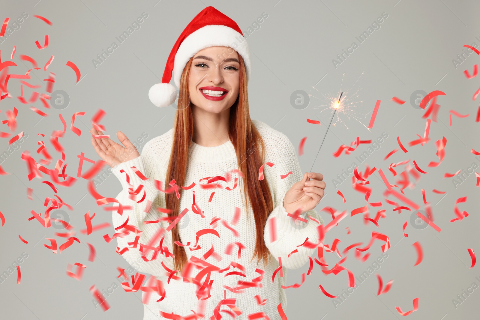 Image of Happy woman in Santa hat with sparkler on grey background. Red confetti flying near her