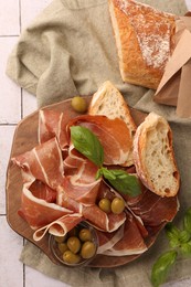 Slices of tasty cured ham, olives, bread and basil on tiled table, flat lay