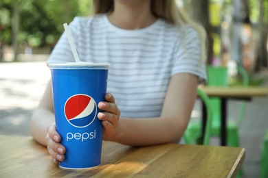 Photo of MYKOLAIV, UKRAINE - JUNE 9, 2021: Woman with paper Pepsi cup at wooden table in outdoor cafe, closeup