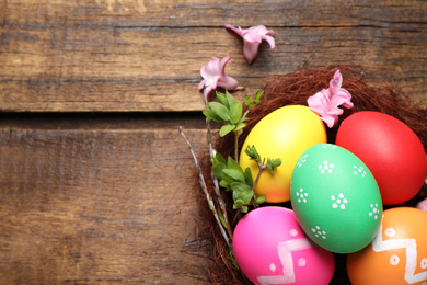 Colorful Easter eggs in decorative nest with flowers on wooden background, flat lay. Space for text