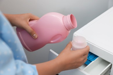 Photo of Woman pouring laundry detergent from bottle into cap near washing machine, closeup