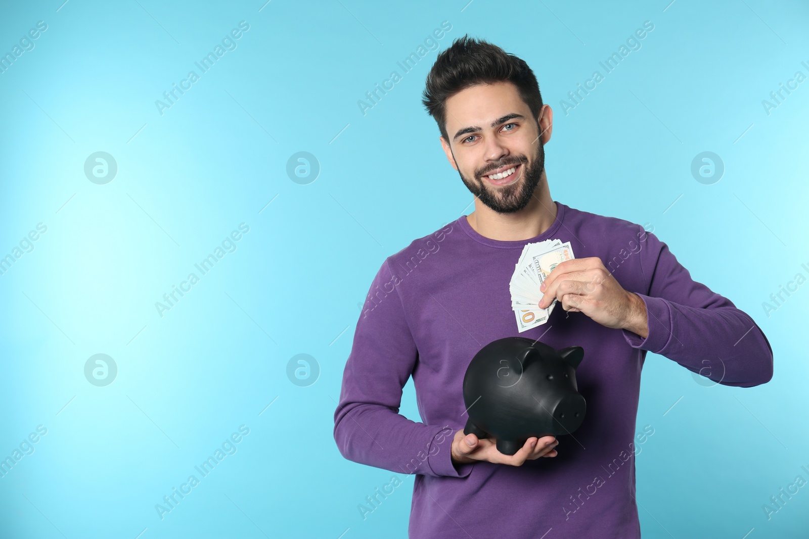 Photo of Happy young man putting money into piggy bank on color background. Space for text
