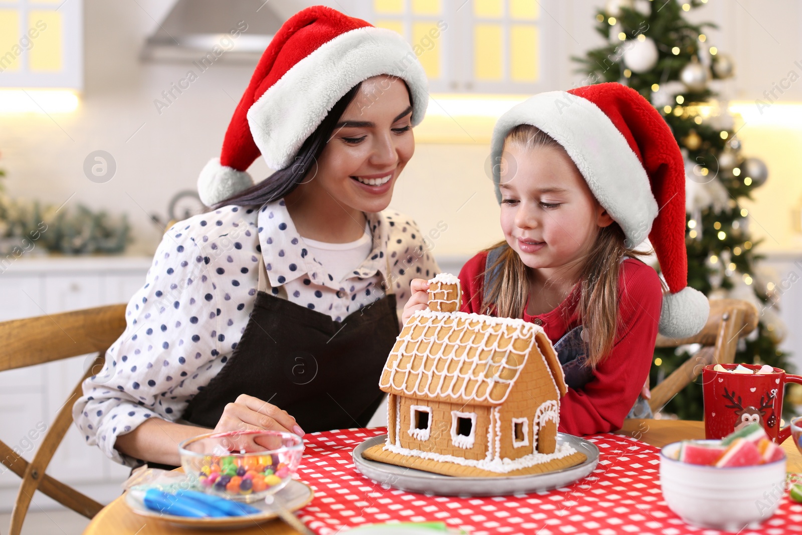 Photo of Mother and daughter decorating gingerbread house at table indoors