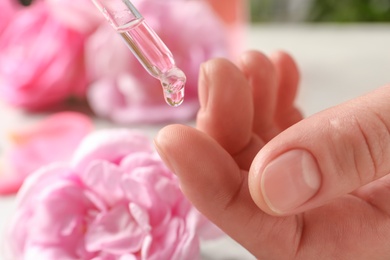 Photo of Woman dripping rose essential oil on finger against blurred flowers, closeup