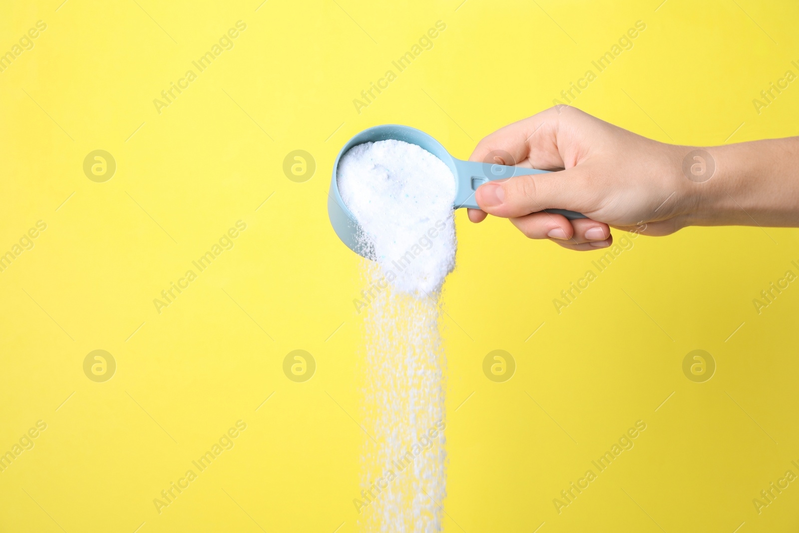 Photo of Woman pouring laundry powder from measuring scoop on yellow background, closeup