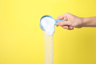 Photo of Woman pouring laundry powder from measuring scoop on yellow background, closeup