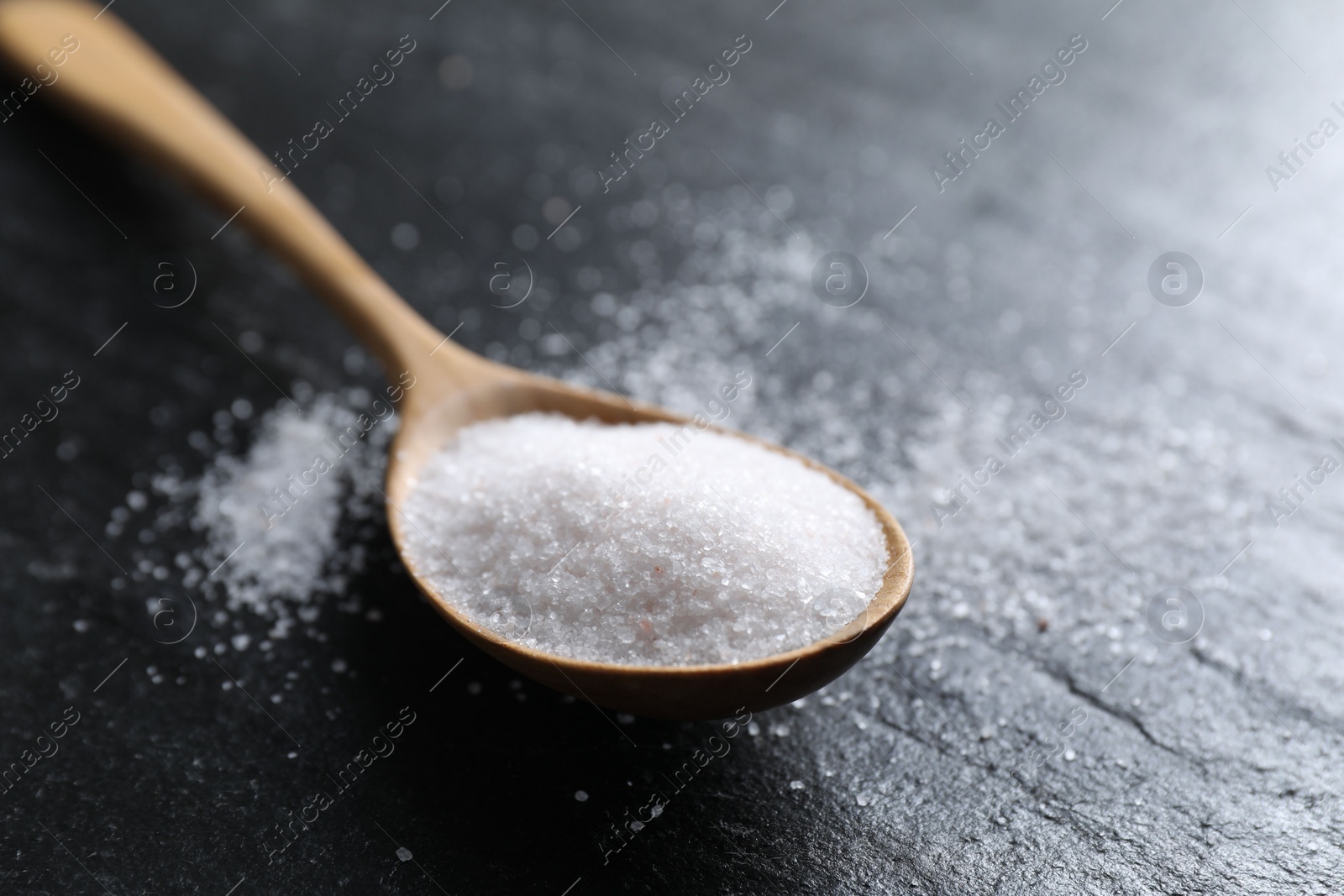 Photo of Organic white salt in spoon on black table, closeup. Space for text