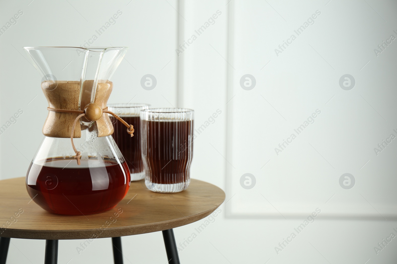 Photo of Glass chemex coffeemaker and glasses of coffee on wooden table against white wall, space for text