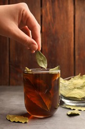 Woman putting bay leaf into cup of freshly brewed tea at grey table, closeup