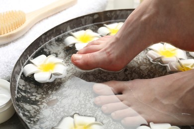 Photo of Woman soaking her feet in bowl with water and flowers on floor, closeup. Spa treatment