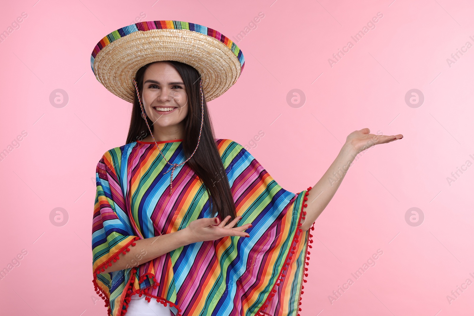 Photo of Young woman in Mexican sombrero hat and poncho showing something on pink background