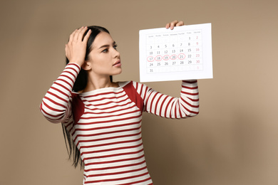 Photo of Emotional young woman holding calendar with marked menstrual cycle days on beige background