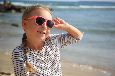 Happy little girl in stylish sunglasses on beach near sea