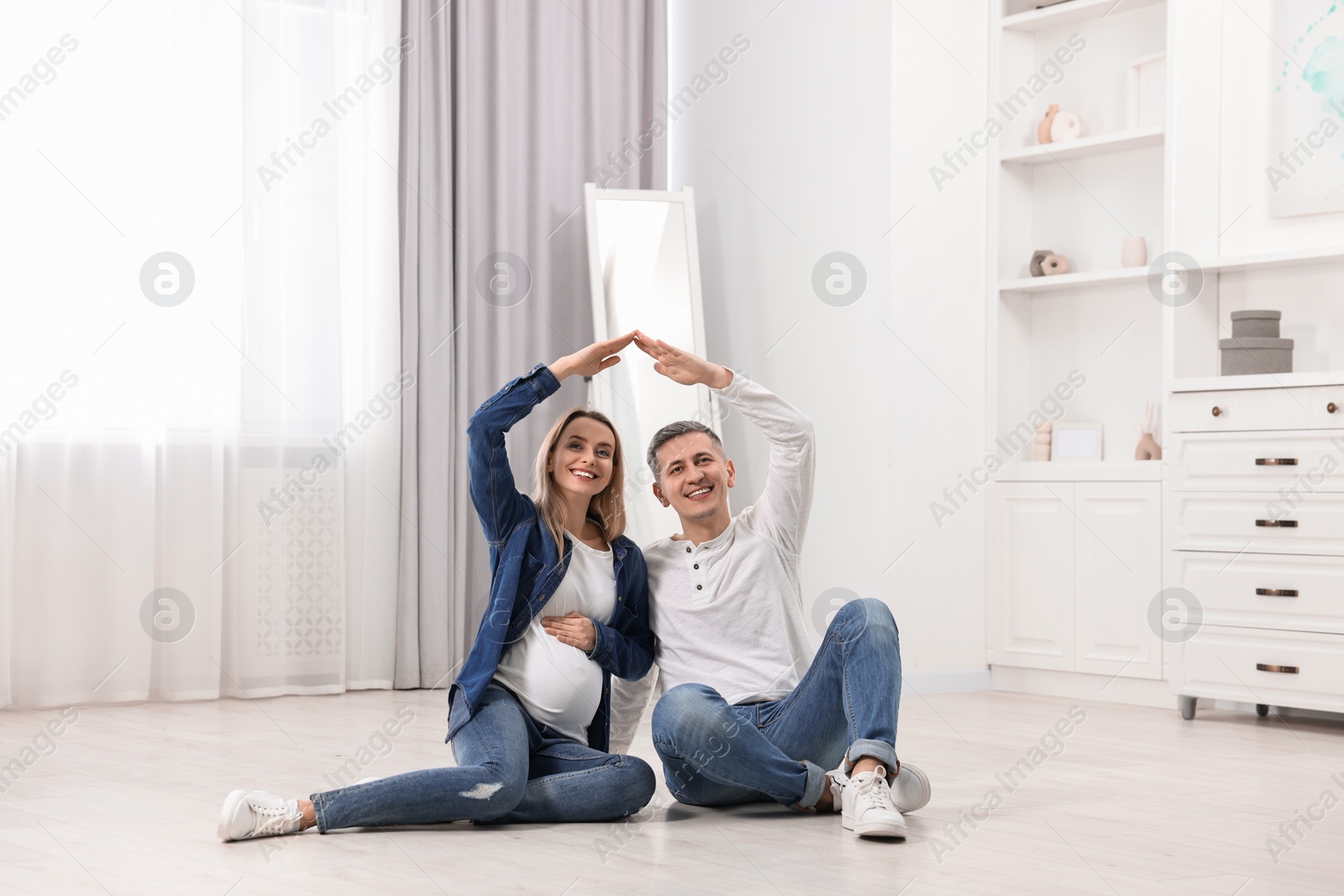 Photo of Young family housing concept. Pregnant woman with her husband forming roof with their hands while sitting on floor at home