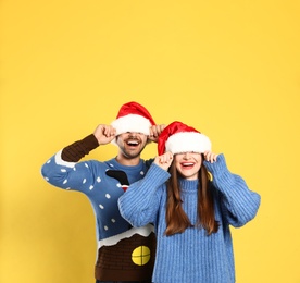 Couple wearing Christmas sweaters and Santa hats on yellow background