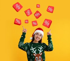 Discount offer. Happy young woman in Christmas sweater and Santa hat pointing at percent signs on orange background