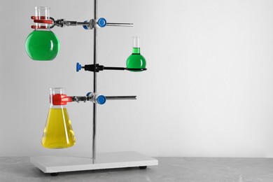 Retort stand and laboratory flasks with liquids on grey table against white background, space for text