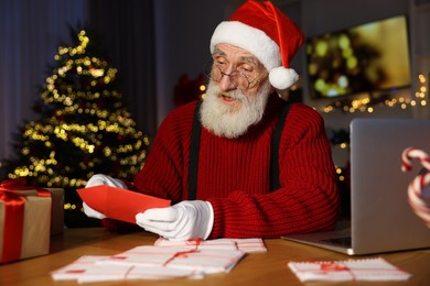 Santa Claus opening letter at his workplace in room decorated for Christmas