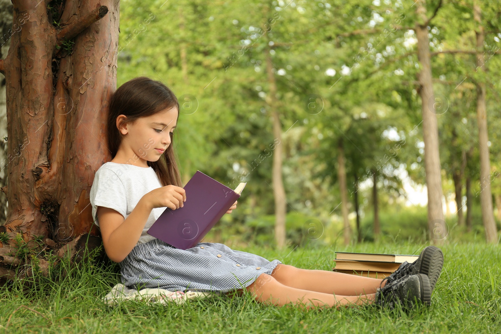 Photo of Cute little girl reading book on green grass near tree in park