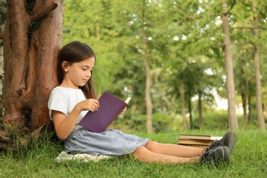 Photo of Cute little girl reading book on green grass near tree in park