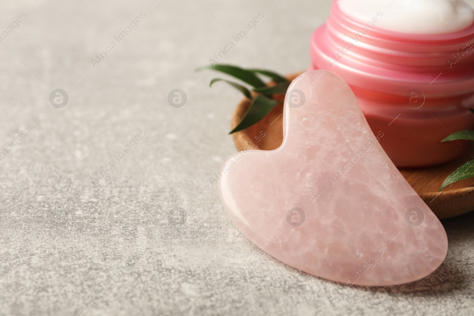 Photo of Rose quartz gua sha tool and jar of cream on grey table, closeup. Space for text